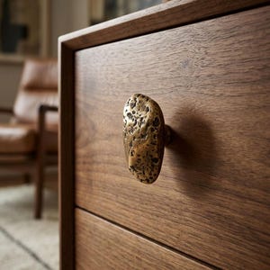 Peut inclure: Une façade de tiroir en bois brun foncé dotée d'un bouton de meuble unique. Le bouton est fabriqué dans un matériau métallique avec une surface texturée, irrégulière et piquée, rappelant une pierre naturelle ou un galet doré ou laiton.