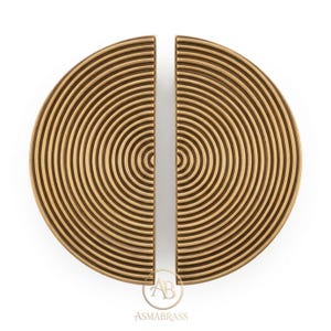 Puede incluir: Dos tiradores de puerta semicirculares de latón con un patrón de nervaduras concéntricas. El acabado metálico es de tono dorado mate. Un logotipo circular con las letras AB y el texto ASMABRASS es visible en la parte inferior central.