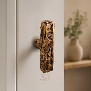 May include: A vertical T-shaped cabinet handle with a textured, rugged, dark gold finish. The handle features a rough, uneven surface and is mounted on a white cabinet door. The logo ASMABRASS is visible in small text at the bottom of the handle.