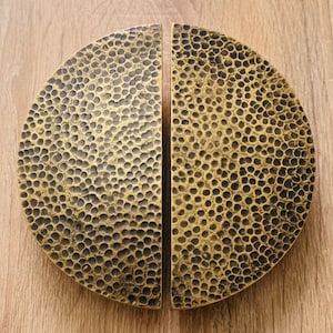 May include: Two semi-circular cabinet pulls made of brass with a hammered, textured surface. Each piece features a dark, antiqued finish within the indentations and a polished gold-tone finish on the raised areas, forming a complete circle.