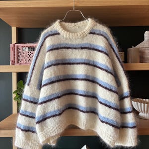 May include: A fuzzy, long-sleeved knit sweater featuring a horizontal striped pattern in cream, light blue, and dark brown. The garment has a round ribbed neckline, dropped shoulders, and ribbed cuffs, crafted from a soft, textured yarn material.