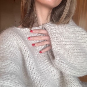 Könnte beinhalten: Ein grob gestrickter Pullover in hellem Beige mit einer dicken, strukturierten Garnkonstruktion, langen Ärmeln und einem weiten Ausschnitt. Das Kleidungsstück hat ein weiches, flauschiges Aussehen mit sichtbaren, großflächigen Strickmaschen.