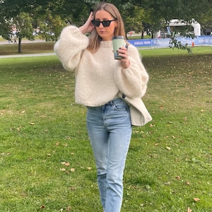 Puede incluir: Un suéter de manga larga esponjoso de color crema con cuello redondo, jeans de mezclilla azul claro, zapatillas de lona blancas, gafas de sol rectangulares negras y un bolso de hombro claro, sosteniendo un vaso de café verde.