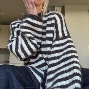 May include: A chunky knit jumper featuring a horizontal striped pattern in dark brown and off-white. The garment has a crew neckline, long sleeves with dropped shoulders, and a relaxed, oversized fit with a fuzzy, textured yarn construction.