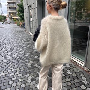 May include: An oversized, cream-colored fuzzy knit sweater with long sleeves and a crew neckline, paired with loose-fitting, light beige trousers. The sweater has a soft, textured appearance and a relaxed fit, while the trousers are smooth and straight-legged.