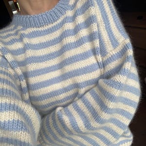 Peut inclure: Un pull en tricot duveteux à manches longues avec des rayures horizontales bleu clair et crème. Le vêtement présente un col rond côtelé et une silhouette décontractée et ample avec des épaules tombantes, fabriqué en fil doux et texturé.