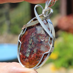 May include: A handcrafted pendant featuring a polished, oval-shaped stone with a mix of red, green, and brown hues. The stone is encased in a delicate silver wire wrap, creating a unique and artistic piece of jewelry.