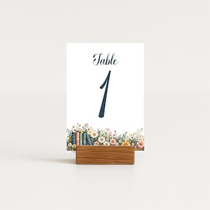 May include: A rectangular white table number card labeled Table 1 in dark blue script and serif font. The bottom features a colorful illustration of small flowers, green foliage, and a row of miniature books, held by a rectangular wooden stand.