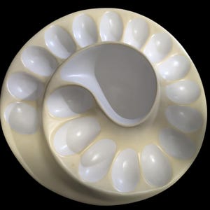 May include: A circular, cream-colored ceramic tray featuring a spiral arrangement of numerous oval-shaped indentations. The surface has a smooth, glossy finish with white-toned concave wells designed for holding small items in a radial pattern.