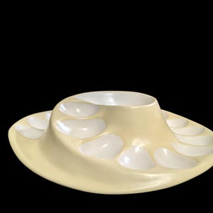 May include: A cream-colored ceramic serving tray featuring a central raised bowl surrounded by a spiral arrangement of twelve individual recessed compartments, each with a smooth white interior finish and a curved, flowing outer edge design.