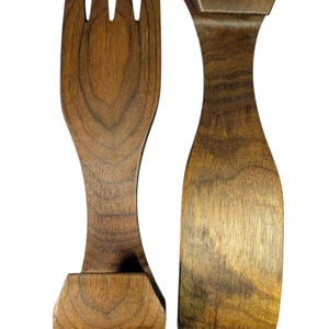 May include: A pair of dark brown wooden salad servers featuring a natural wood grain pattern. One server is shaped like a fork with three tines, while the other is a solid, curved paddle. Both utensils have a smooth finish and a wide, flat handle base.