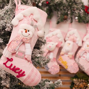 Puede incluir: Un calcetín navideño rosa claro con una aplicación de muñeco de nieve de felpa, una bufanda de lentejuelas y un detalle de techo de tela blanca. El nombre Lucy está bordado en cursiva rosa oscuro y la puntera tiene un patrón de cuadros.