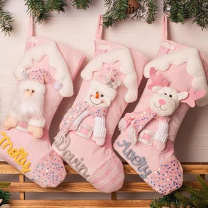 May include: Three light pink Christmas stockings featuring plush white trim at the top. Each stocking displays a different character: a Santa Claus, a snowman, and a reindeer. The names Melly, Devin, and Avery are embroidered on the stockings.