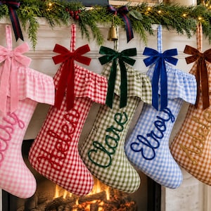 Puede incluir: Cinco calcetines navideños colgantes con patrones de cuadros vichy en rosa, rojo, verde, azul y tostado. Cada calcetín presenta un puño con volantes, un lazo de terciopelo y un nombre bordado en cursiva: Betsy, Helen, Clare, Elroy y Adele.