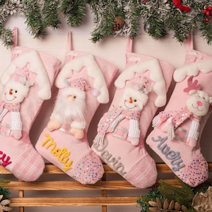 Op de afbeelding: Vier lichtroze kerstsokken met witte pluche boorden en geborduurde namen. Elke sok toont een uniek figuur: twee sneeuwpoppen, een kerstman en een rendier, allemaal voorzien van bijpassende roze sjaals en decoratieve accenten.