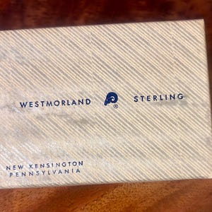 May include: A rectangular off-white box featuring a diagonal striped pattern. The text reads WESTMORLAND, a ram logo, and STERLING in the centre, with NEW KENSINGTON PENNSYLVANIA printed in the bottom left corner in dark blue lettering.