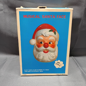 May include: A rectangular box featuring a Musical Santa Face with a red hat, white beard, and a red nose. The blue packaging includes the text Musical Santa Face, Plays Santa Claus Is Coming To Town, and Turn Nose To Activate Music.