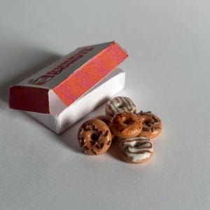 May include: A miniature white and orange box labeled Dunkin Donuts containing a small pile of tiny, realistic-looking toy donuts. The donuts feature various glazes, including chocolate with sprinkles and white icing swirls, crafted in a small scale.