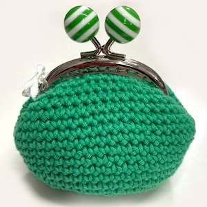 Può includere: Un portamonete all'uncinetto color verde menta con una chiusura a scatto in metallo argentato. La chiusura è sormontata da due perline sferiche decorate con strisce diagonali bianche e verdi alternate. Un piccolo nastro bianco è fissato sul lato.