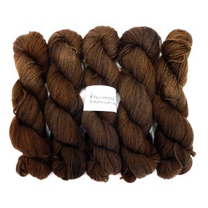 May include: Five skeins of brown yarn, in a rich, dark shade, are displayed against a white backdrop. The yarn's colour has subtle variations. A white label with the text "FOLKESTONE & HARBOUR YARNS" is visible.