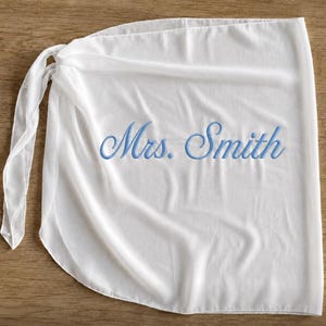 May include: A white wrap skirt featuring a tie closure on the left side. The fabric is lightweight and semi-sheer. Embroidered in the center in a light blue cursive script is the text Mrs. Smith, which is clearly visible against the white background.
