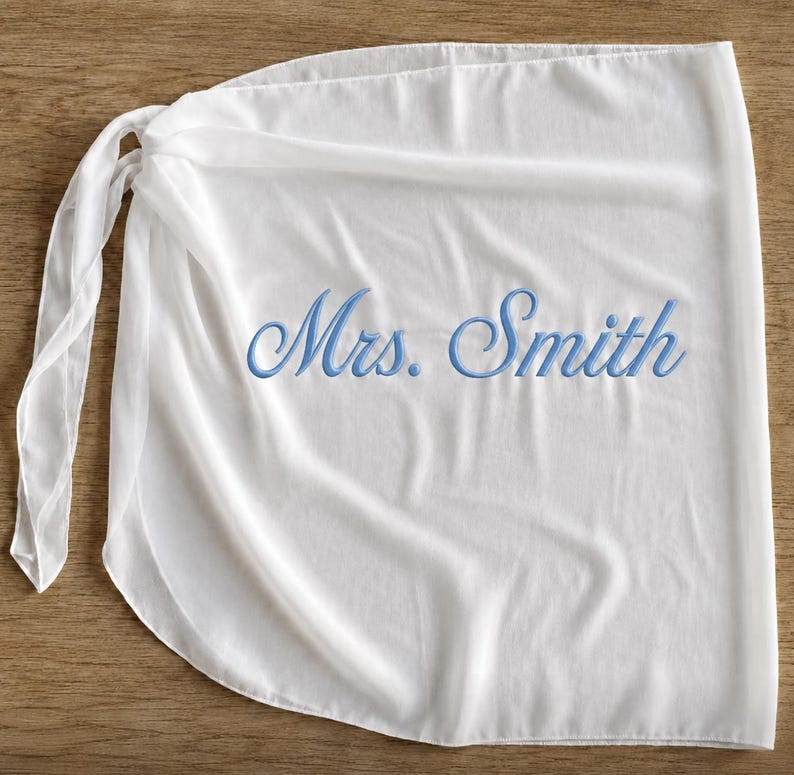 May include: A white wrap skirt featuring a tie closure on the left side. The fabric is lightweight and semi-sheer. Embroidered in the center in a light blue cursive script is the text Mrs. Smith, which is clearly visible against the white background.