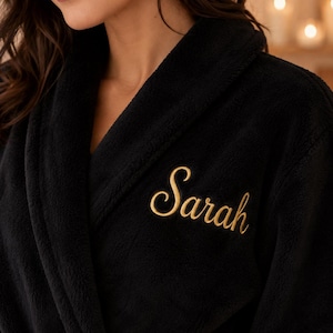 May include: A black plush bathrobe featuring a shawl collar and long sleeves. The left chest area is embroidered with the name Sarah in a gold-colored cursive script font. The fabric has a soft, textured appearance throughout the garment.