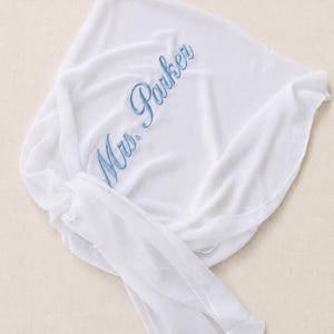 May include: A white sheer fabric wrap featuring the embroidered text Mrs. Parker in a light blue script font. The lightweight material is draped with a knotted tie detail on one side, showcasing a smooth texture and soft folds throughout the piece.