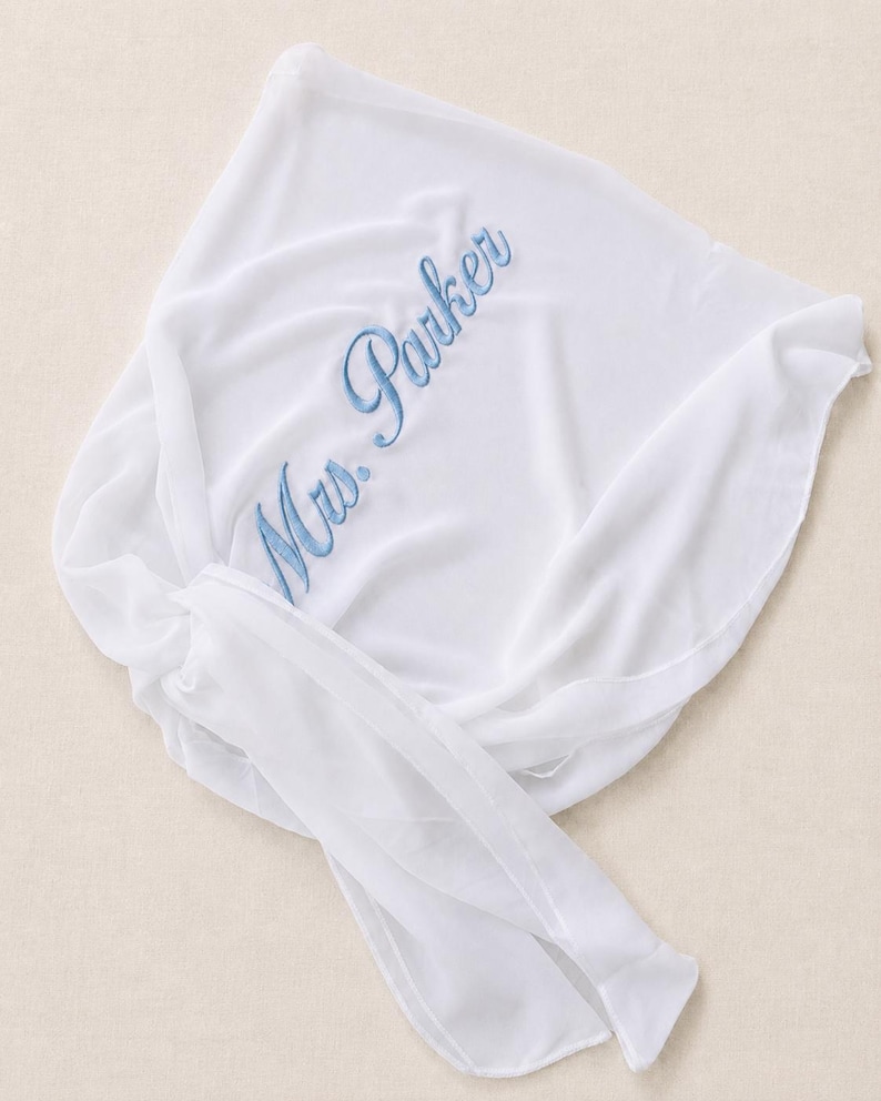 May include: A white sheer fabric wrap featuring the embroidered text Mrs. Parker in a light blue script font. The lightweight material is draped with a knotted tie detail on one side, showcasing a smooth texture and soft folds throughout the piece.