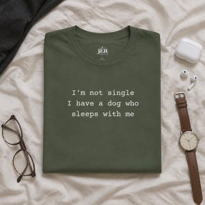 May include: An olive green crew neck t-shirt featuring the white text "I'm not single I have a dog who sleeps with me" printed on the chest. The shirt has a round neckline and short sleeves, with the brand name "JER Factory" printed inside the collar.