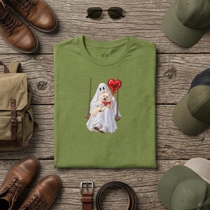 May include: An olive green t-shirt featuring a graphic of a white ghost sitting on a wooden swing, holding a small white dog with a red bandana and a shiny red heart-shaped balloon. The shirt has a crew neckline and the text JER printed inside.