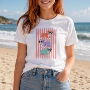 May include: A white short-sleeved t-shirt featuring a graphic print of three puppies wearing sunglasses, stacked vertically against a background of vertical pink stripes. The puppies are colored orange, purple, and teal, each wearing dark sunglasses.
