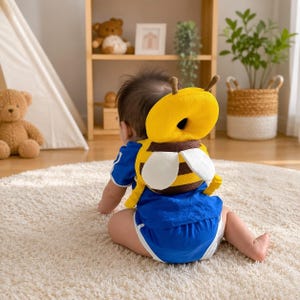 May include: A baby head protector cushion shaped like a bumblebee, featuring a bright yellow circular head pad, brown and yellow striped body section, and two white wings. The accessory includes two small antennae and is worn like a backpack.