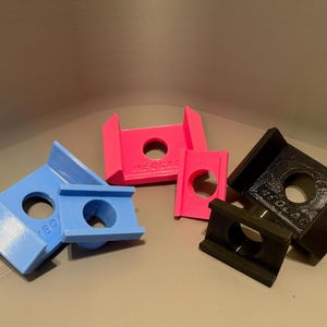 May include: A collection of six U-shaped plastic brackets in light blue, bright pink, and black. Each bracket features a central circular hole and the embossed text "RISOCAD" on the side. The items are arranged in pairs of varying sizes.