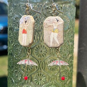 May include: A pair of dangle earrings featuring rectangular ceramic tiles with illustrations of figures in traditional attire. One figure holds a cloth, the other a bow. Below each tile hangs a miniature translucent umbrella charm with red heart accents.