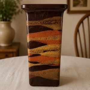 May include: A tall, rectangular ceramic vase featuring a horizontal striped pattern in shades of dark brown, burnt orange, and mustard yellow. The surface has a distinct crackle glaze texture throughout, with a smooth, dark brown rim at the top.