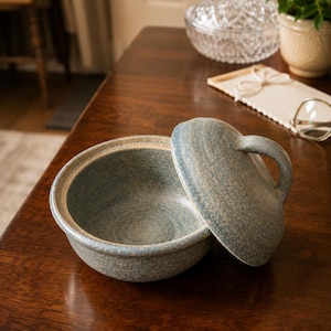 May include: A round, speckled blue ceramic bowl with a matching lid. The bowl has a wide, open interior and a smooth, curved rim. The lid features a curved handle on top, and both pieces have a textured, mottled finish in shades of blue and tan.