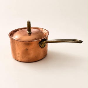 May include: A round copper saucepan with a matching lid and a long brass handle. The pot features a polished metallic finish, a vertical brass knob on the center of the lid, and a sturdy brass handle attached to the side of the vessel.