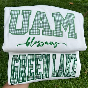 May include: A white sweatshirt featuring the green and white gingham block letters UAM, the cursive text blossoms in green embroidery, and the block letters GREEN LAKE in green and white gingham pattern on the lower section of the garment.