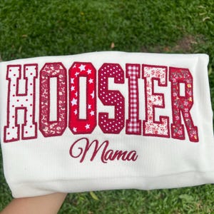 May include: A white sweatshirt featuring the word HOOSIER in large, block-style letters, each filled with a different red and white pattern including polka dots, florals, stars, and gingham. Below, the word Mama is embroidered in red script.