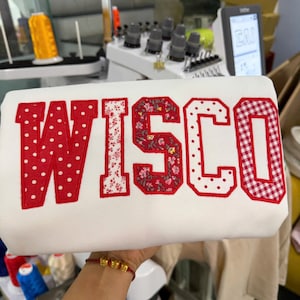 May include: A white sweatshirt featuring the embroidered letters WISCO in a collegiate font. Each letter has a red stitched border and a unique pattern: the W is red with white dots, the I is white with red flowers, the S is red with floral print, the C is white with red dots, and the O is red with a white gingham check pattern.
