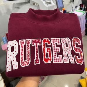 May include: A maroon sweatshirt featuring the word RUTGERS in large, block-style letters. Each letter is filled with a different pattern, including red and white polka dots, floral prints, stars, and a red and white gingham check pattern.