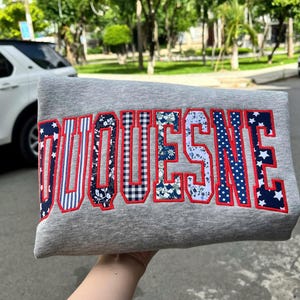 May include: A heather gray sweatshirt featuring the word DUQUESNE appliqued in large block letters. Each letter is filled with a different fabric pattern, including stars, stripes, gingham, and floral prints, all outlined in bright red stitching.