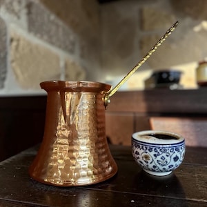 May include: A hammered copper coffee pot with a long, angled brass handle, positioned next to a small, round ceramic cup featuring a blue and white floral pattern filled with dark liquid coffee, reflecting light on its metallic surface.