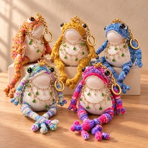 May include: Five plush frog keychains in orange, yellow, blue, multicolored, and pink, each featuring a white belly, black eyes, a small gold crown, and a gold chain. Each frog wears a decorative gold-tone necklace with dangling green crystals.