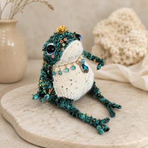 May include: A handcrafted crochet frog figurine featuring a dark teal textured body with iridescent sequins, a cream-colored belly with silver accents, large dark eyes, a small gold crown, and a decorative gold-tone necklace with blue crystals.