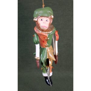 May include: A hanging ornament shaped like a monkey wearing a green beret and a multi-colored jester-style outfit. The costume features orange panels with red star patterns, green striped sleeves, and gold accents. The monkey has a light brown face.