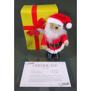 May include: A beige mohair teddy bear dressed as Santa Claus with a red hat, white beard, black belt, and black boots. The bear wears wire-rimmed glasses. It is accompanied by a yellow gift box with a red ribbon and a Steiff certificate of authenticity.
