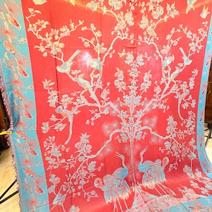 May include: A large rectangular textile featuring a vibrant red background with a light blue border. The fabric displays a detailed pattern of flowering branches, various birds perched on limbs, and two large cranes standing at the base.