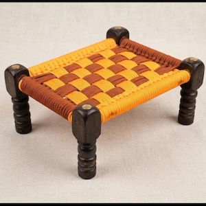 May include: A small rectangular stool featuring four dark brown turned wooden legs with brass-toned circular accents on top. The seat is constructed from a woven checkerboard pattern of bright yellow and brown cord, stretched across the wooden frame.